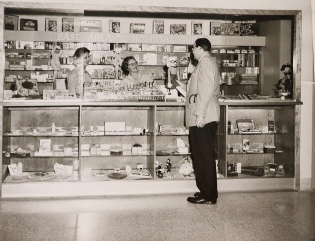 Two women stand behind the counter at the gift shop while a man shops.