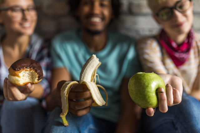 Three people holding something different, a donut, a banana and an apple