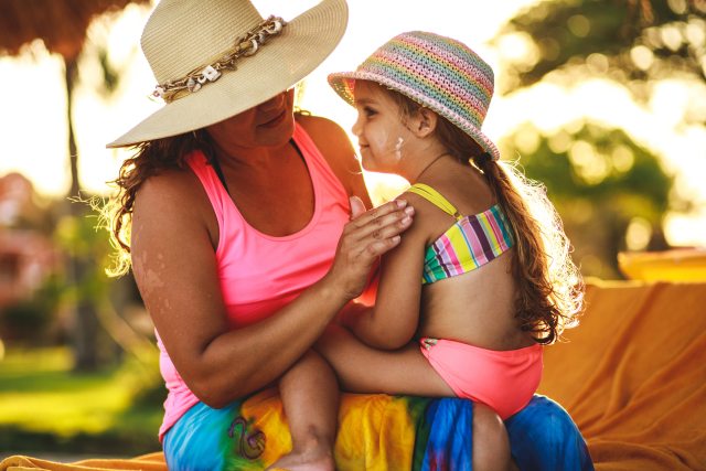 A mother sitting on a beach chair wearing a sun hat applies sunscreen to her young daughter's shoulder.