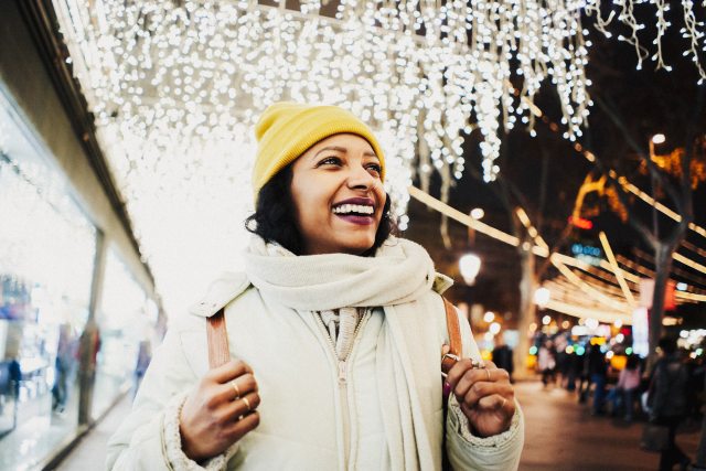 Woman in yellow hat and white scarf smiles outside brightly lit stores