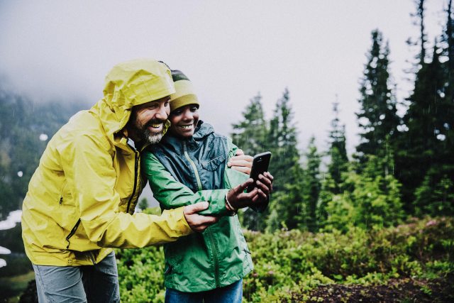 A man and boy in rain jackets take a selfie while on a hike in the rainy forest.