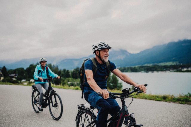 An older couple ride bicycles down a concrete path near a body of water