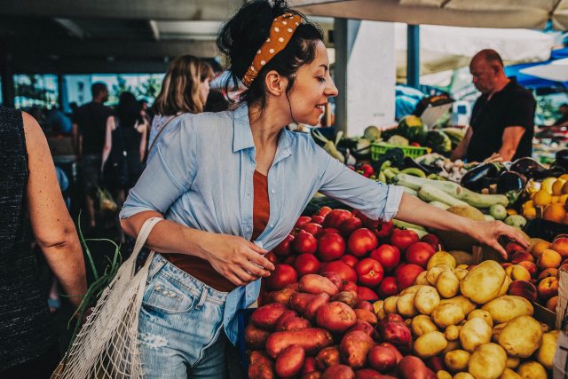 A woman at a farmer's market reaches over a vegetable stand to grab a potato