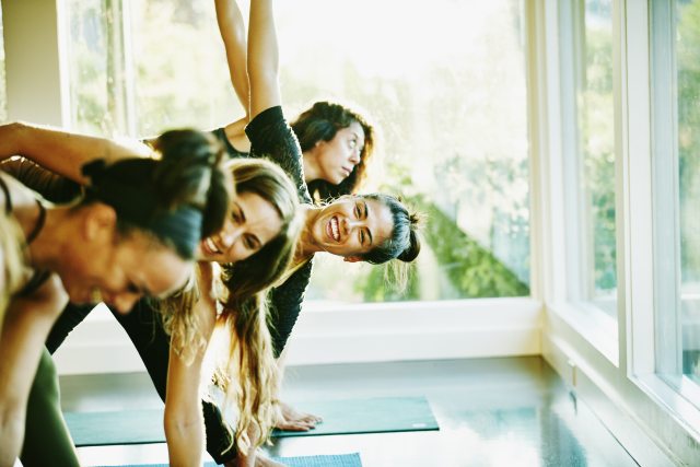 A group of women in a well lit room, laugh and smile during a yoga session