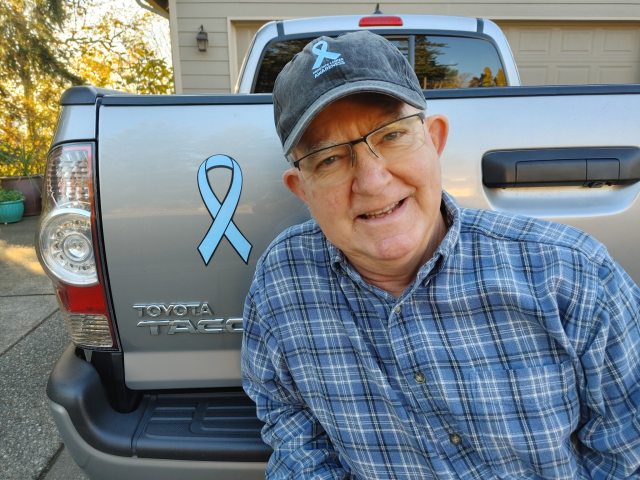 Skip Gibson smiling next to a blue cancer ribbon sticker on the back of a truck