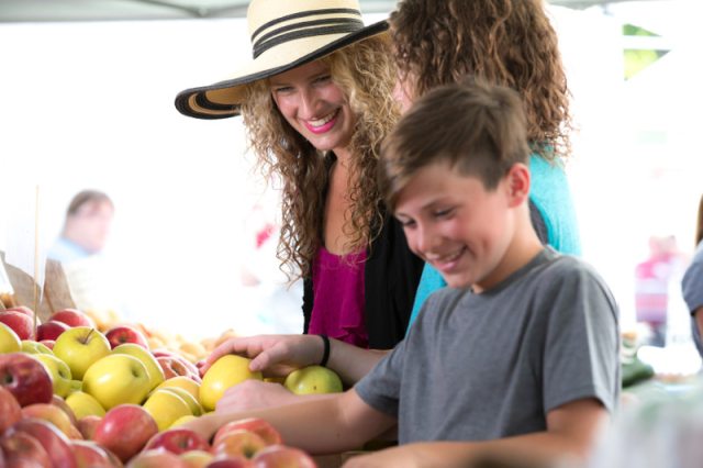 Three people smile and look through vegetables at a farm stand