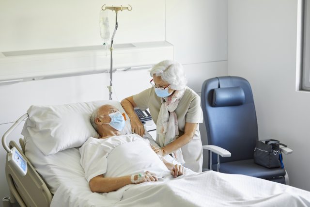 An older patient lies in bed while being comforted by an elderly visitor