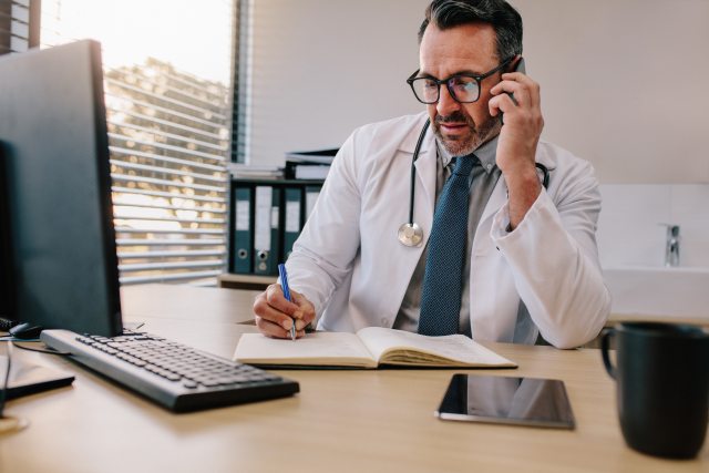 A healthcare provider at a desk and computer writes in a journal while speaking on the phone