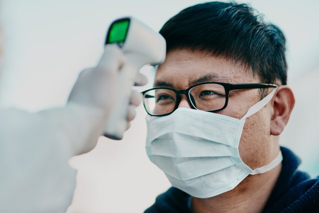A patient in a mask has their forehead scanned for a temperature reading