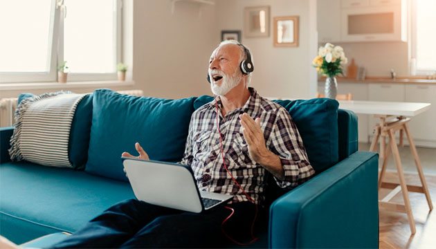A man sitting on a couch with a laptop and headphones, singing or reacting jubilantly to the sound