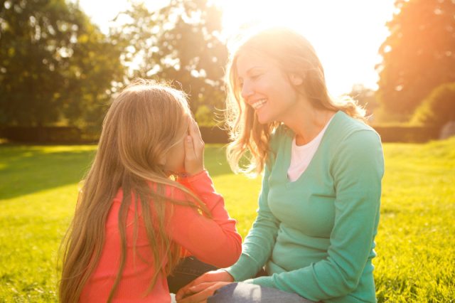 Mother and daughter playing peek-a-boo while sitting in a park, with sublight in the background
