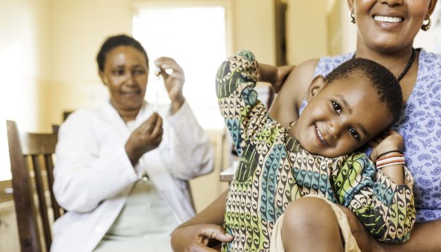 A child smiles in the foreground, with a health care provider in the background
