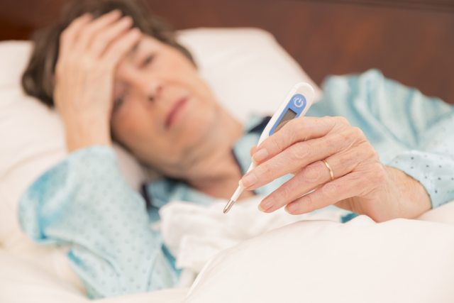 An older lady laying down takes her tempaerature and rests hand on her forehead.