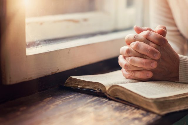Folded hands in prayer resting on a bible next to the window