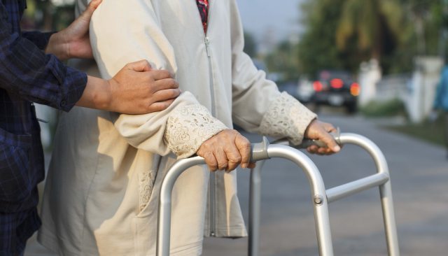 An older person walks across the street with a walker and the aid of someone off camera