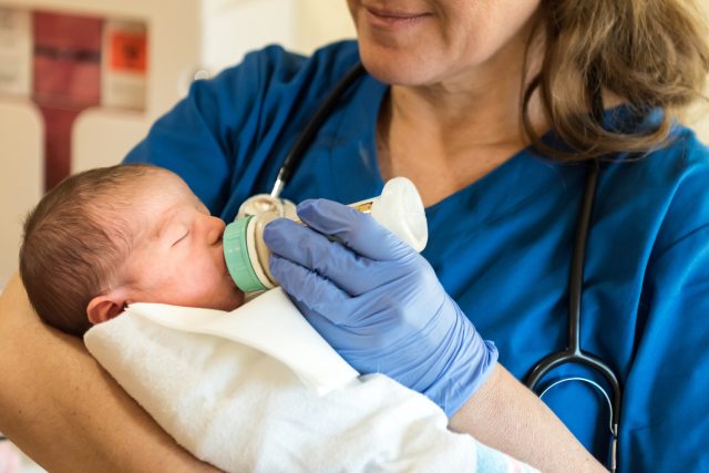 A gloved nurse in scrubs smiles and feeds a newborn baby with a bottle