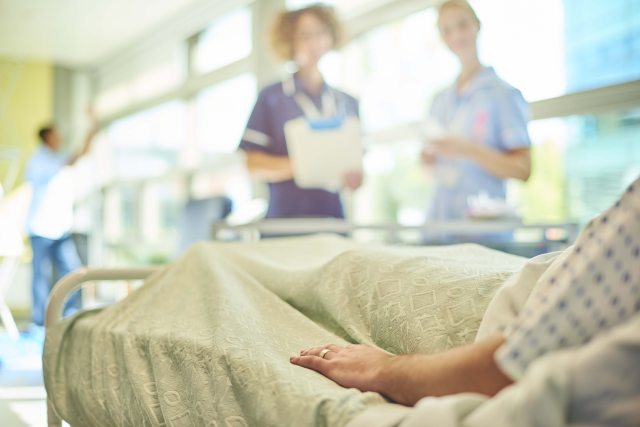 2 Healthcare providers with charts stand in the background, while a patient lays on a bed in the foreground