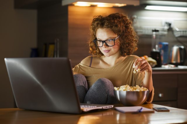 A young adult watches a laptop in a dimly lit kitchen, with popcorn on her left