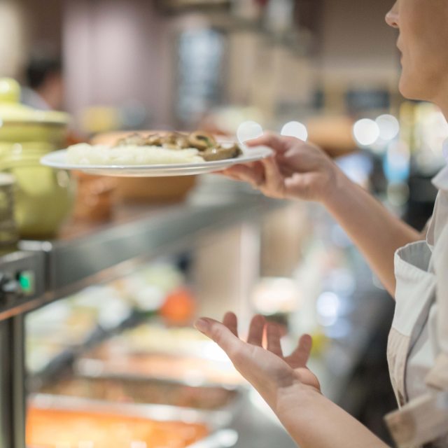 A person collects a plate of food in the cafeteria lunch line