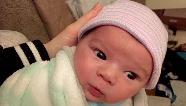 A wide-eyed newborn baby with a pink and blue striped beanie being cradled by an adult